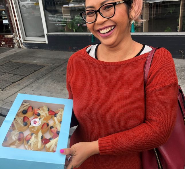 Smiling woman holding a box of cupcakes outside.